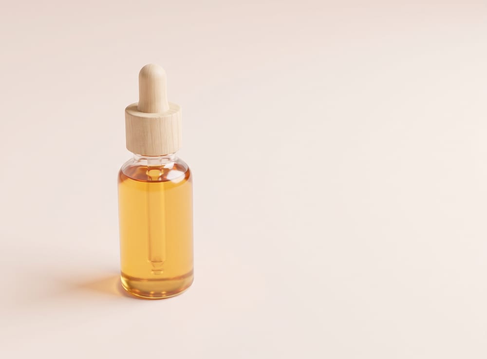 A small clear glass bottle of golden hair oil with a wooden dropper cap.
