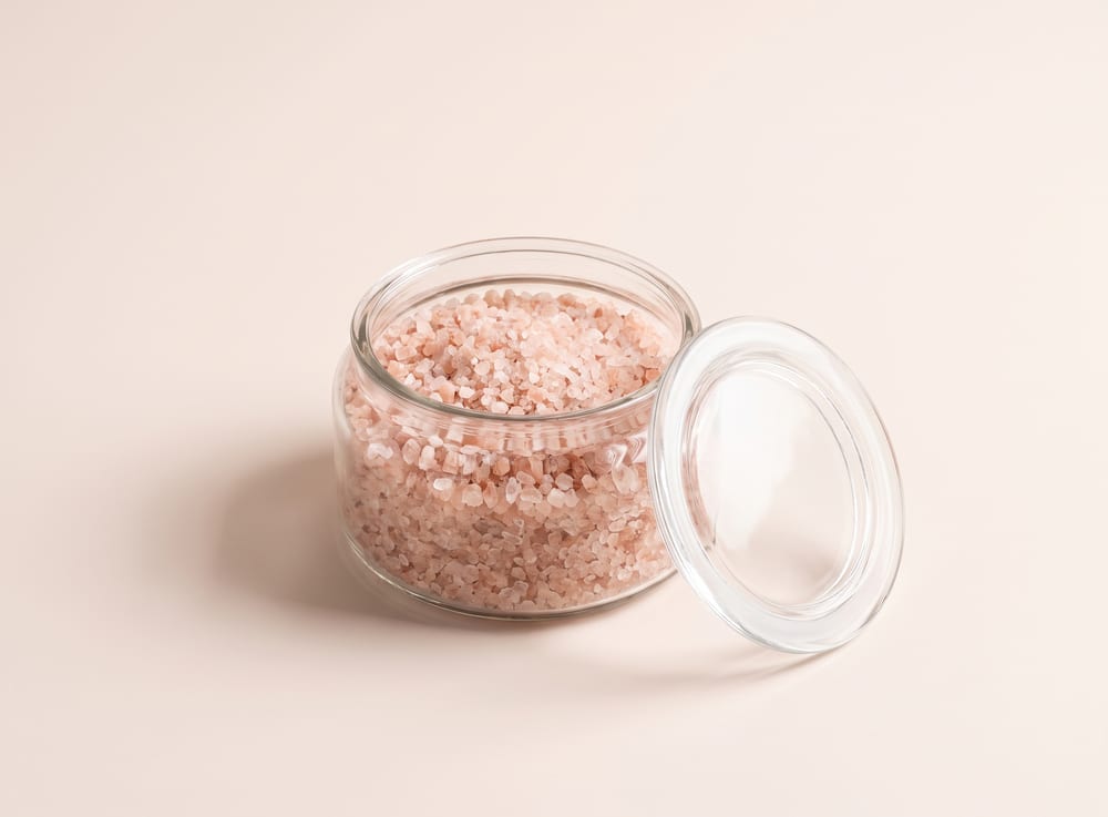 A shallow glass jar of pale pink bath soak crystals with the lid resting beside it.
