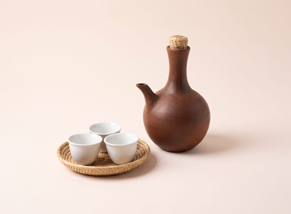 A round unglazed clay coffee pot with a long neck, beside small handleless cups on a woven tray.